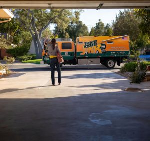 Hyperrealistic 16k image of same day junk removal in Sacramento with TCJ Hauling truck (dark green #02423D with orange #FEA104 stripes) pulling away, a relieved Sacramento homeowner waving goodbye, clean empty garage, crisp daylight, oak trees